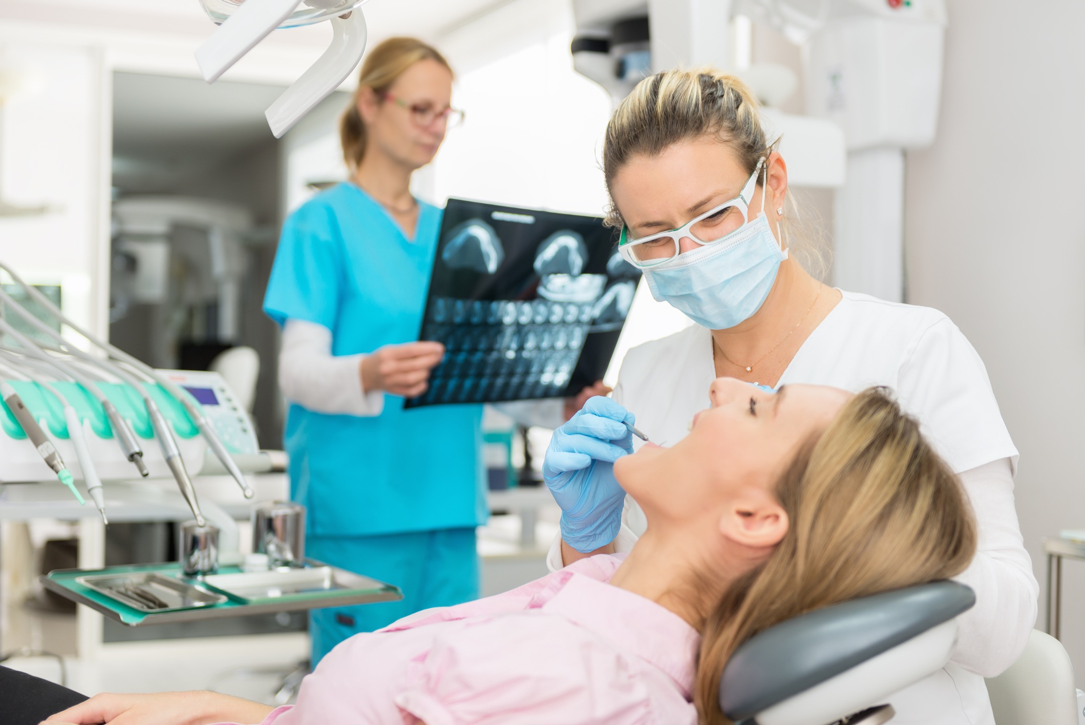 Horizontal color image of three people - dentist, dental assistant and patient at dentist's office. Beautiful young woman having dental examination. Dental assistant standing in background and analyzing x-ray.